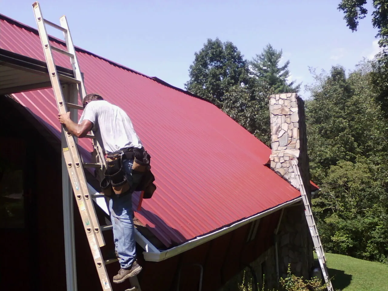 Roofer on a ladder beside a red metal roof and stone chimney — Soffit & Fascia Repair by Roofing Brothers in Hiawatha